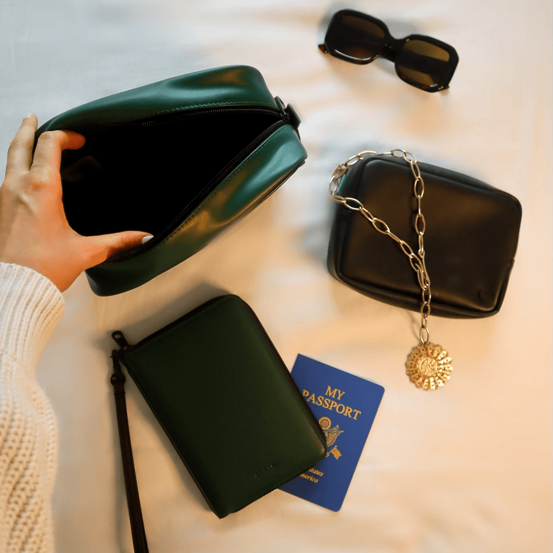 Olive and black pouches styled on a bed with sunglasses, a passport, and a gold necklace
