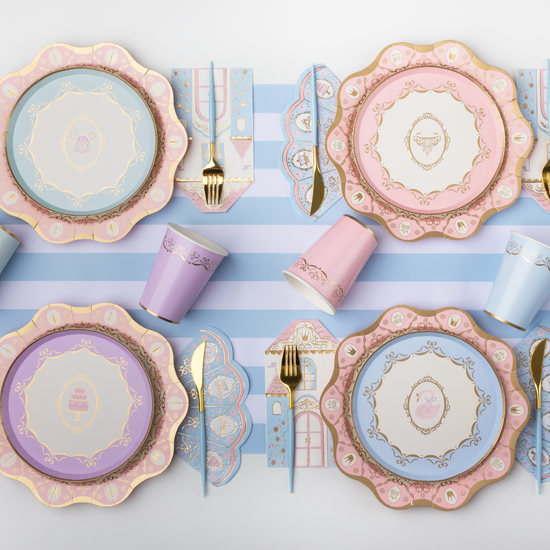 A styled flat-lay of pastel party tableware on a white surface with light blue and white stripes. The set includes scalloped plates in pink, lavender, and light blue with gold detailing, matching cups, napkins, and gold-toned cutlery.
