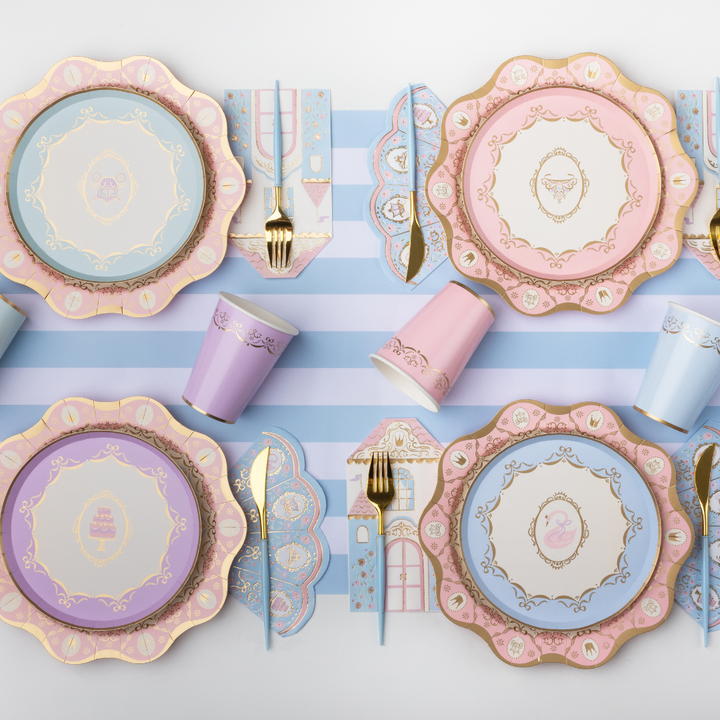 A styled flat-lay of pastel party tableware on a white surface with light blue and white stripes. The set includes scalloped plates in pink, lavender, and light blue with gold detailing, matching cups, napkins, and gold-toned cutlery.