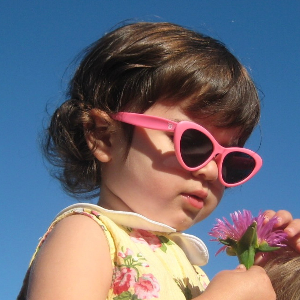 A toddler in a yellow floral dress wearing pink cat-eye sunglasses outdoors and holding a flower under a blue sky.