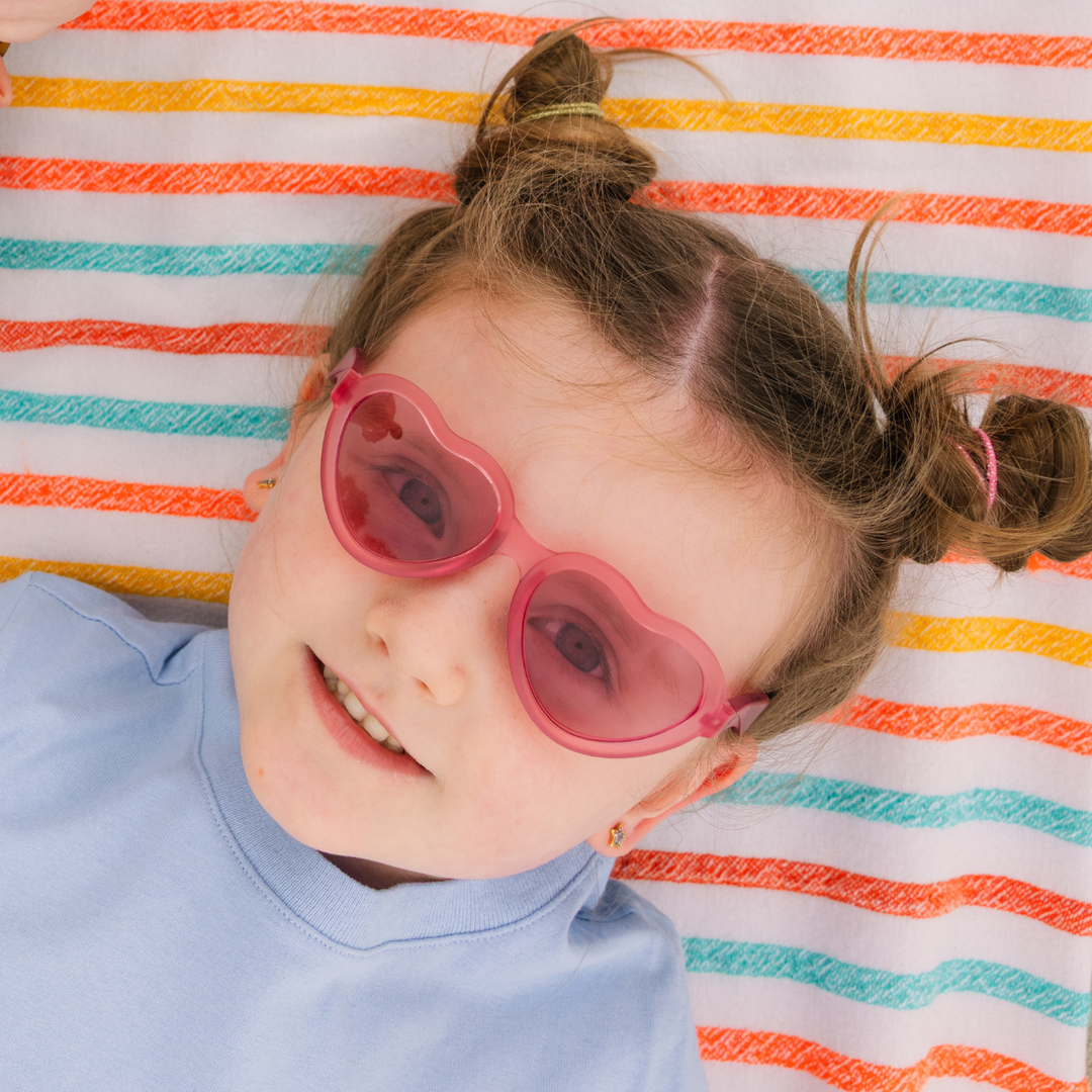 A young girl lying on a striped towel and smiling while wearing fuchsia heart-shaped sunglasses.