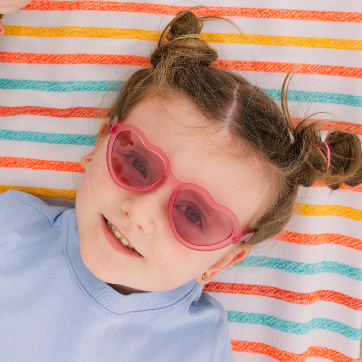 A young girl lying on a striped towel and smiling while wearing fuchsia heart-shaped sunglasses.