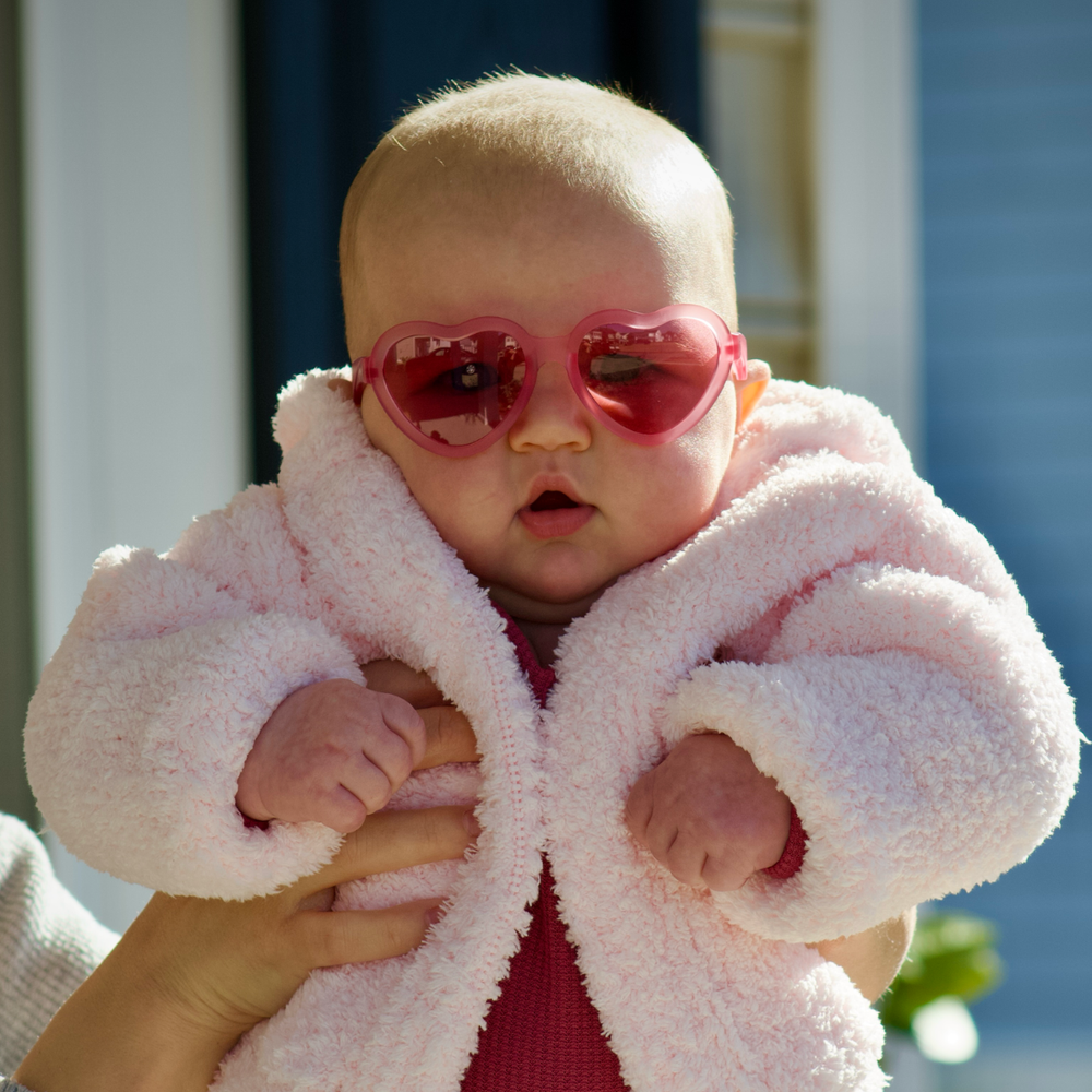 A baby in a pink fleece coat wearing fuchsia heart-shaped sunglasses outdoors on a sunny day.