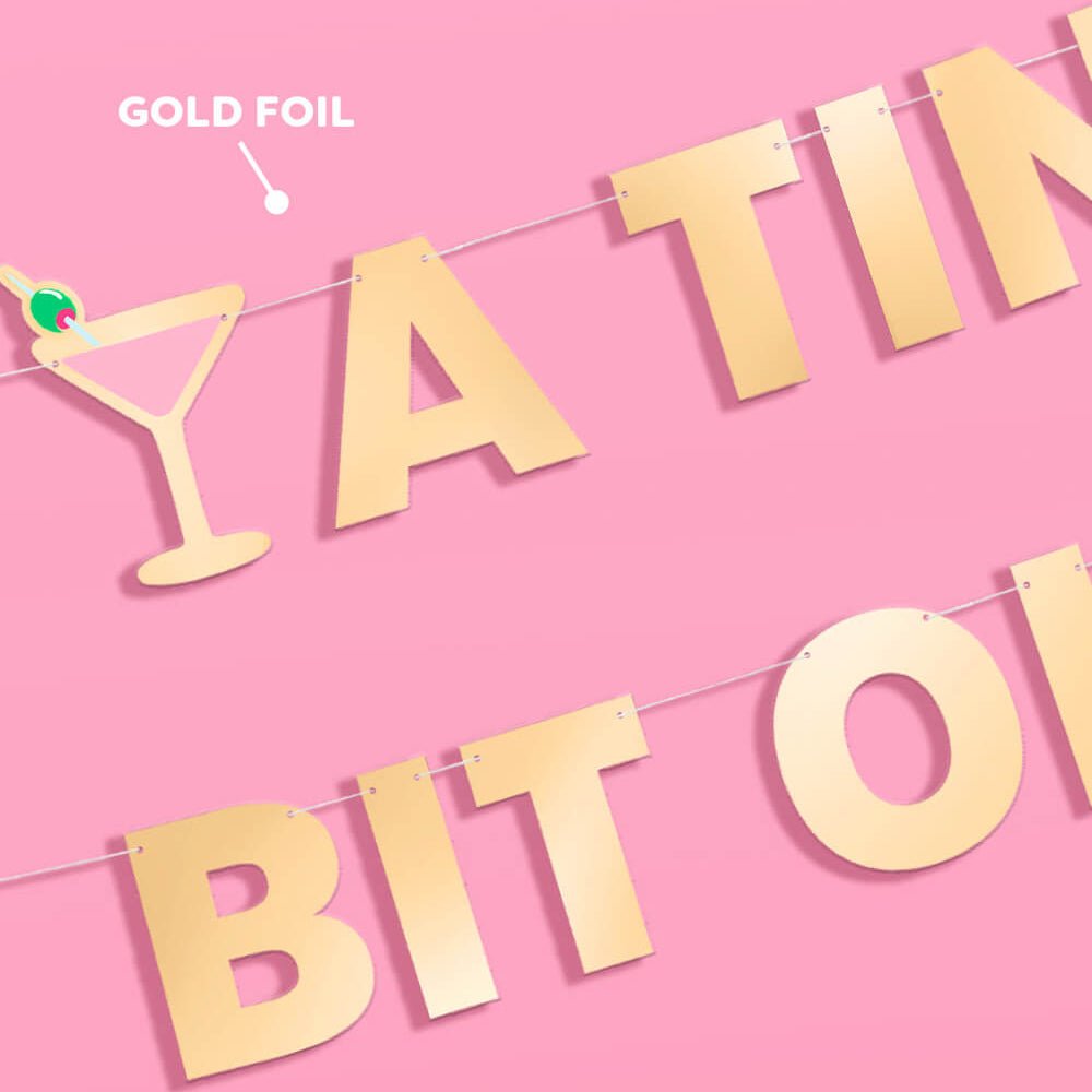 Close-up of gold foil letters and a pink martini glass cutout on the A Tini Bit Older banner, labeled gold foil