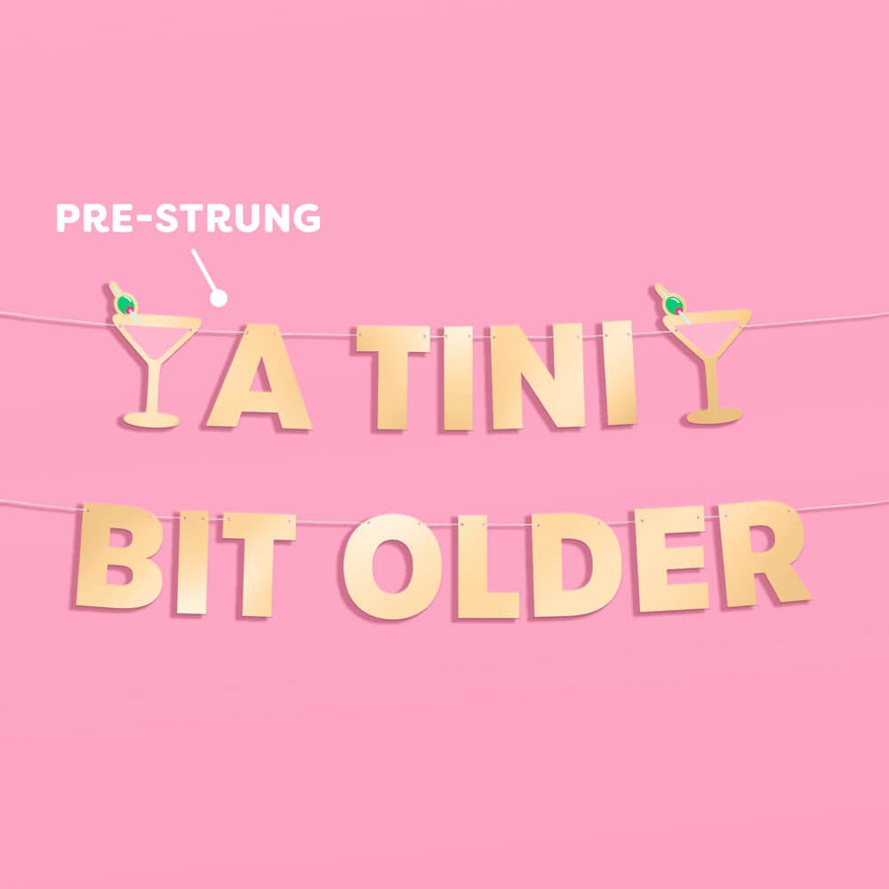 A Tini Bit Older banner on pink showing two pre-strung lines of gold foil letters and pink martini glasses