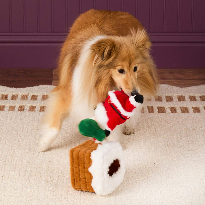 Long-haired dog lifting Santa from the chimney toy while the empty chimney tips onto its side.