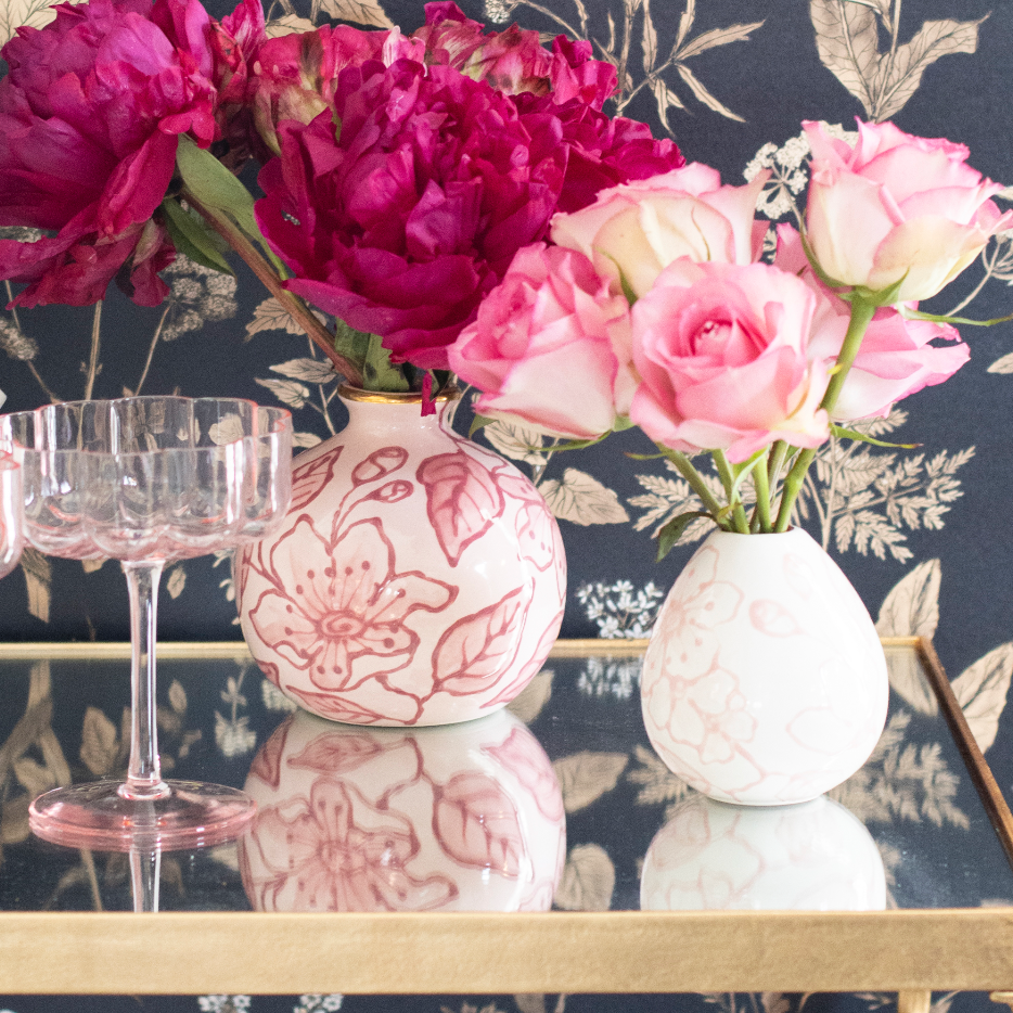 Two pink blossom bud vases with peonies and roses on a mirrored gold bar cart beside a pink coupe glass