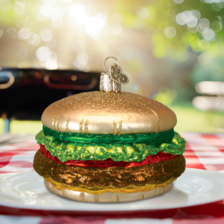 Cheeseburger ornament on a white plate atop a red gingham picnic cloth with a grill behind it