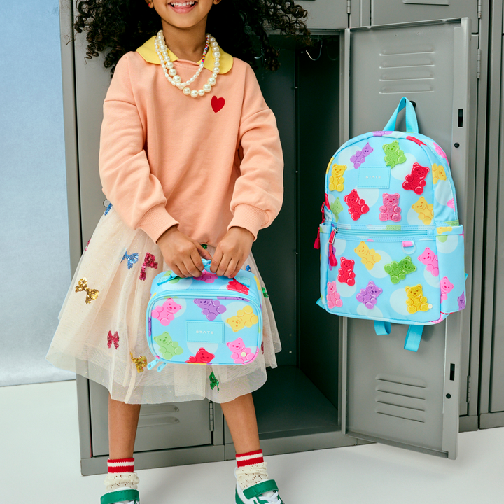 Girl in a tulle skirt by school lockers holding a gummy bear lunch box and backpack