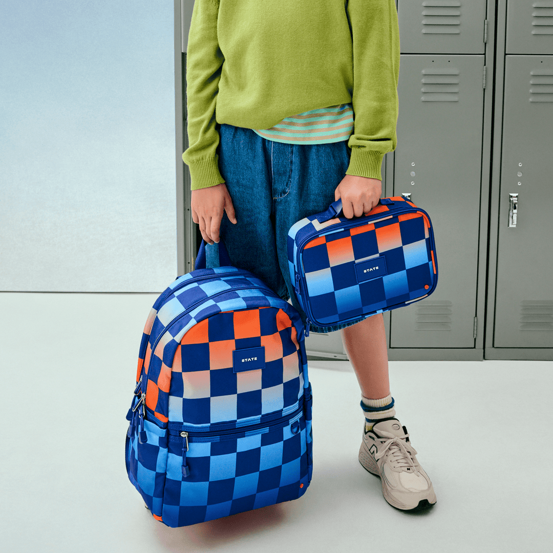 Child by school lockers holding the Gradient Checkerboard lunch box and matching backpack