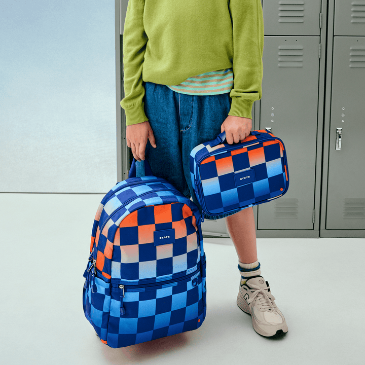 Child by school lockers holding the Gradient Checkerboard lunch box and matching backpack