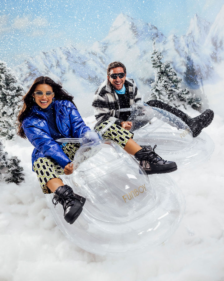 Two riders on a clear inflatable snowmobile sled in the snow in front of snowy mountains
