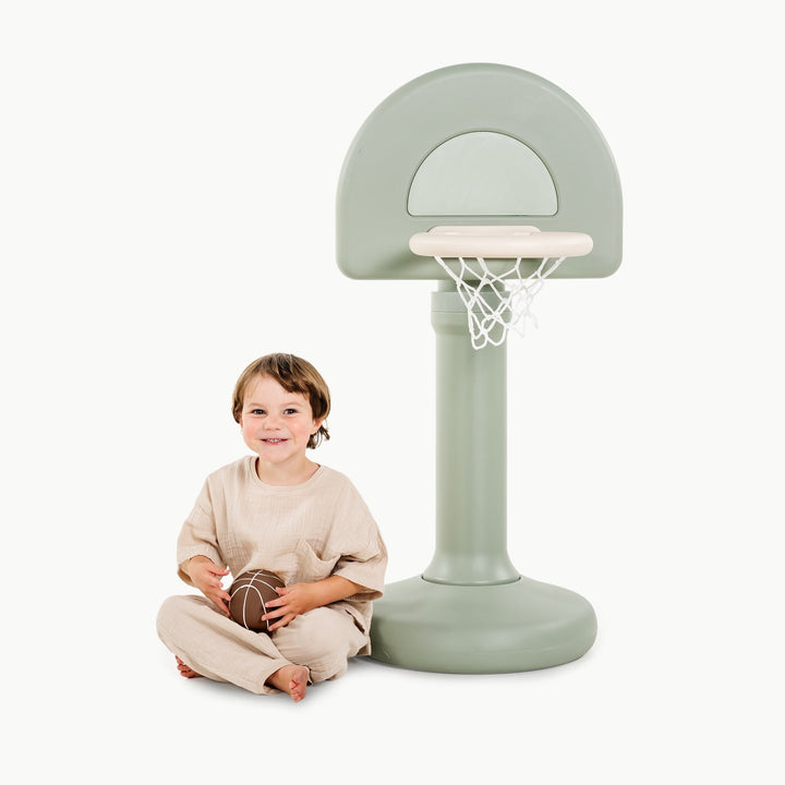 Smiling boy sitting at the base of the palm green hoop holding a basketball