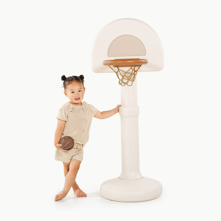 Toddler girl holding a basketball and leaning on the ivory hoop post