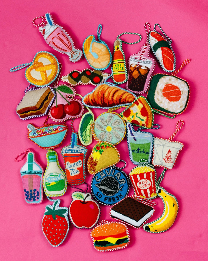 Flat lay of Furbish food needlepoint ornaments on pink, including the PB&J sandwich, burger, and strawberry