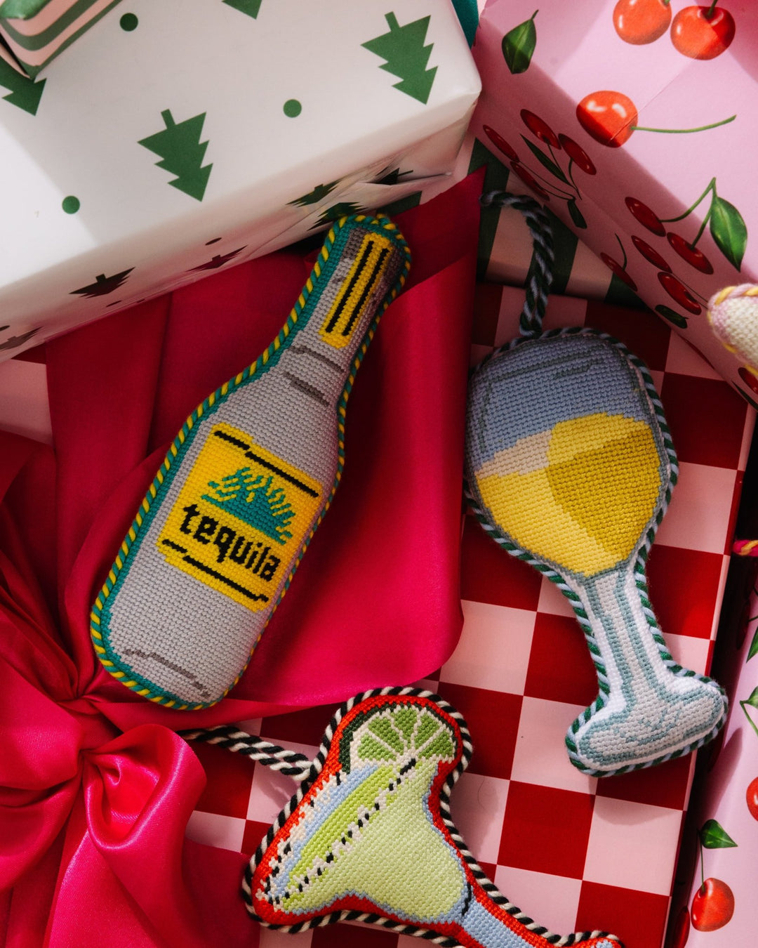 Tequila ornament on a red and checkered gift beside white wine and margarita needlepoint ornaments