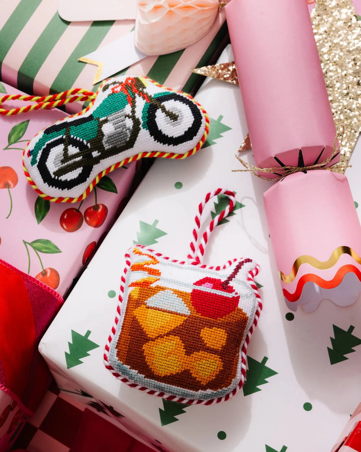 Motorcycle needlepoint ornament on a wrapped gift beside a cocktail ornament and a pink cracker