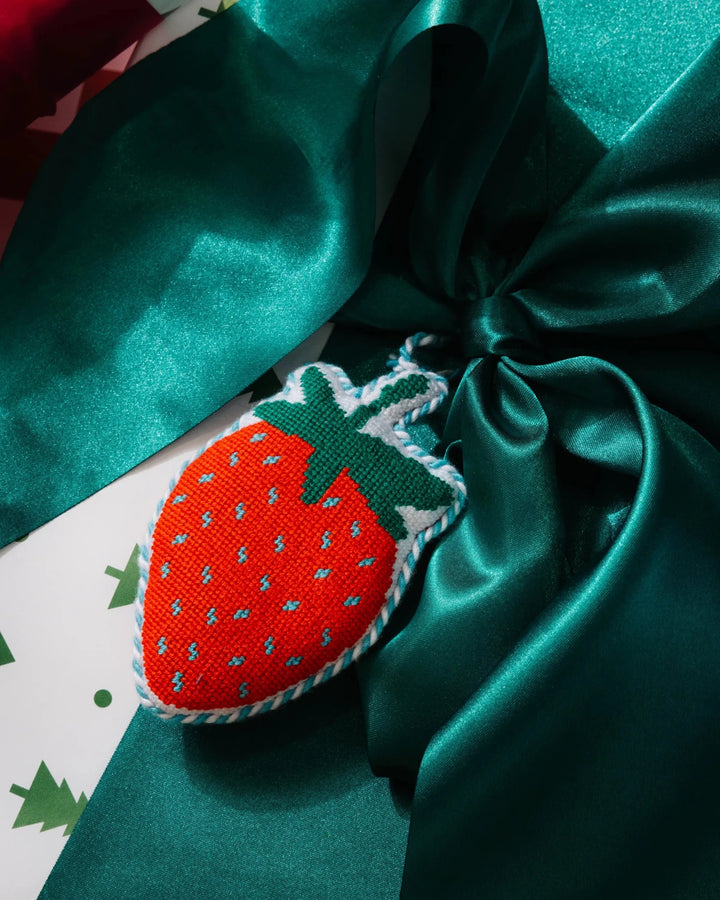 Strawberry needlepoint ornament resting on a green satin bow over tree print wrapping paper