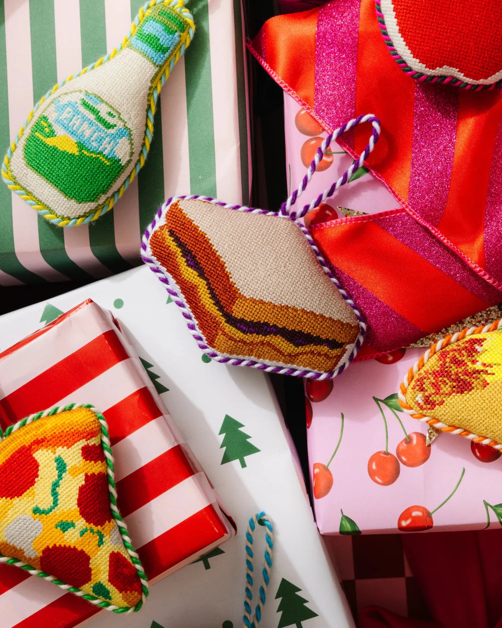 PB&J needlepoint ornament resting on wrapped gifts beside a pizza slice and a soda bottle ornament