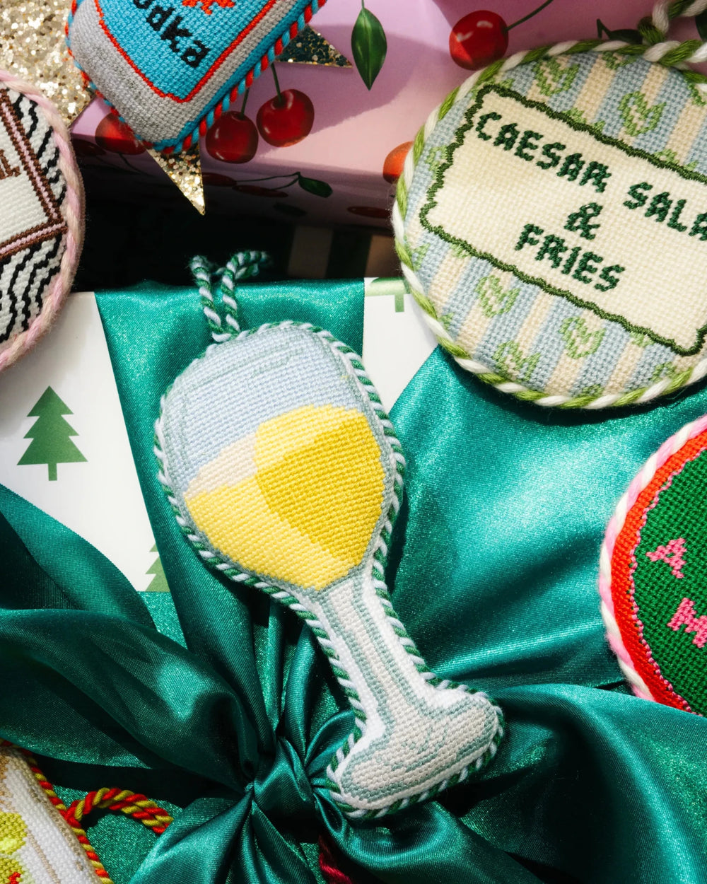 White wine ornament on green satin beside a round Caesar salad and fries needlepoint ornament