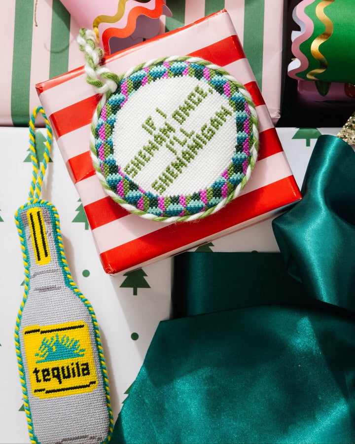 Tequila bottle ornament tucked against a red striped gift box with a round needlepoint saying ornament