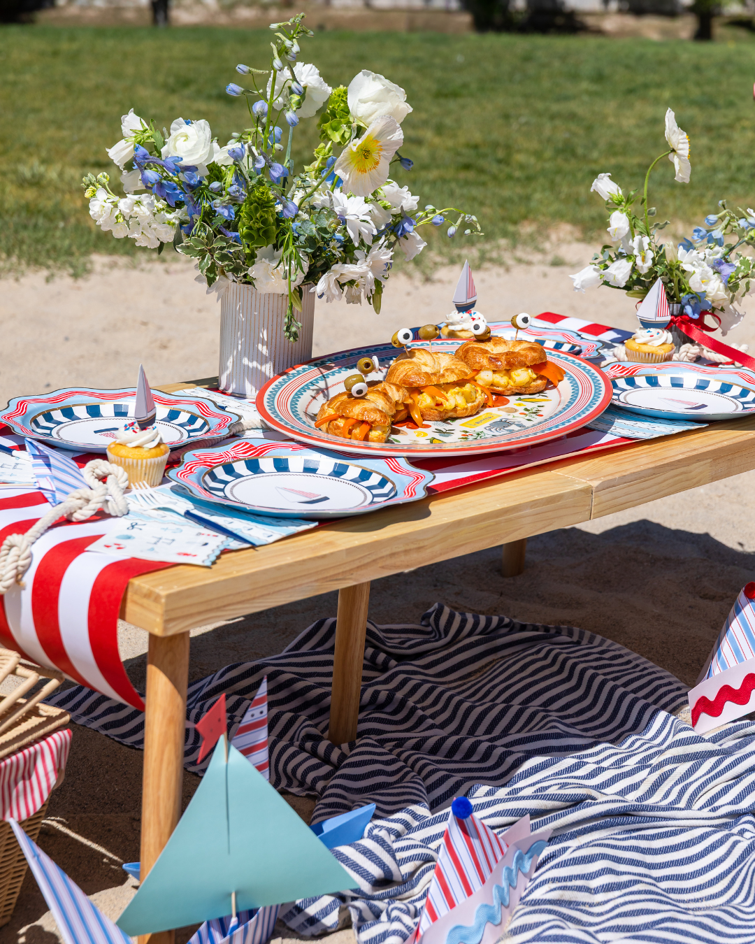 Nautical red, white, and blue beach picnic tablescape with sailboat plates, a striped runner, and croissant sailboat snacks for a summer sailing party