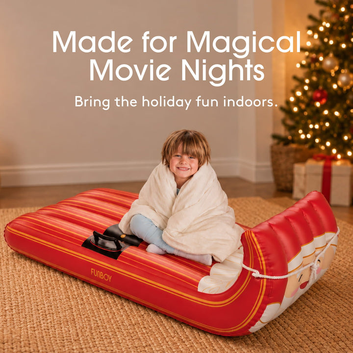 Child wrapped in a blanket sitting in a Santa inflatable sled by a Christmas tree for a movie night