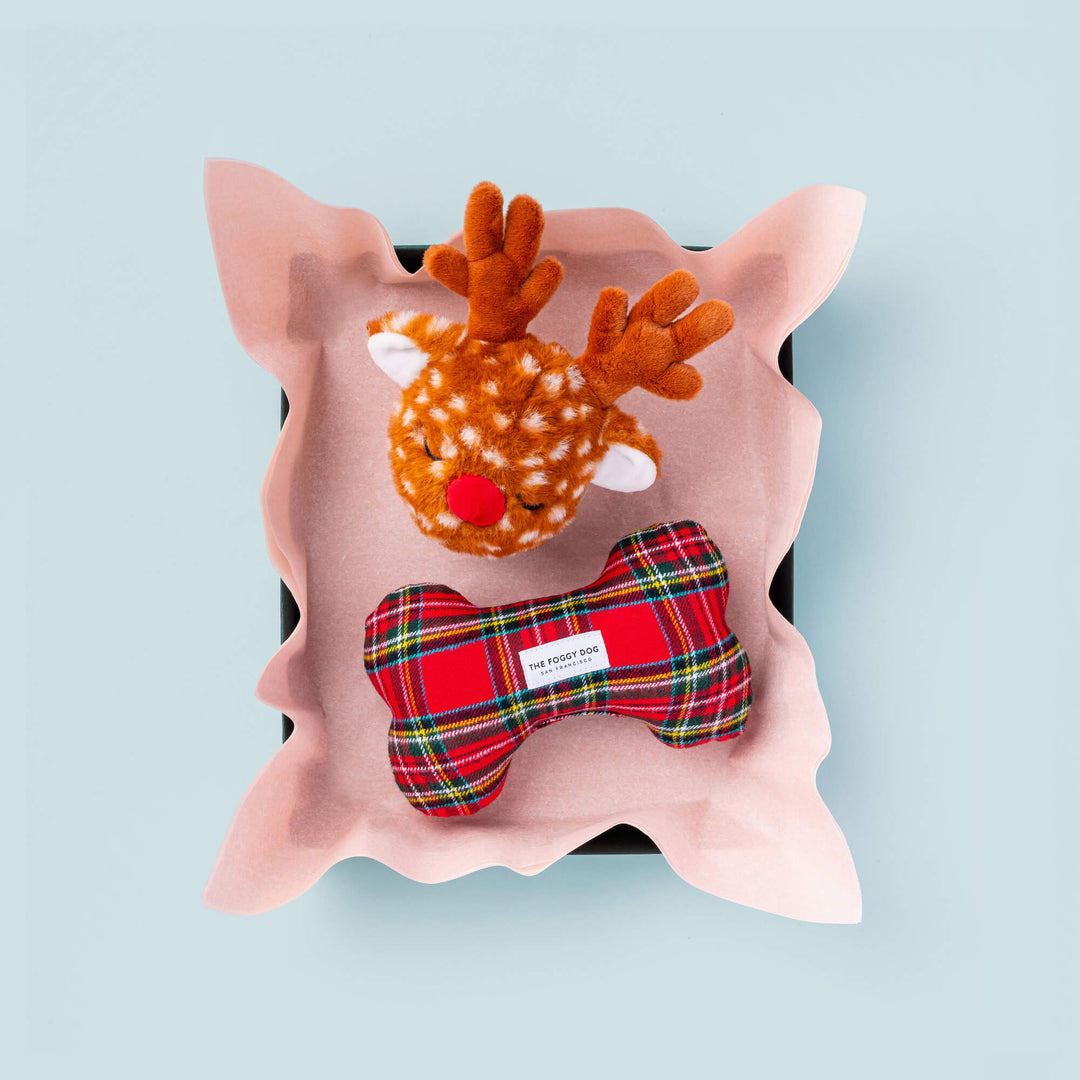 Open box with pink tissue holding the tartan bone toy and a plush reindeer on pale blue