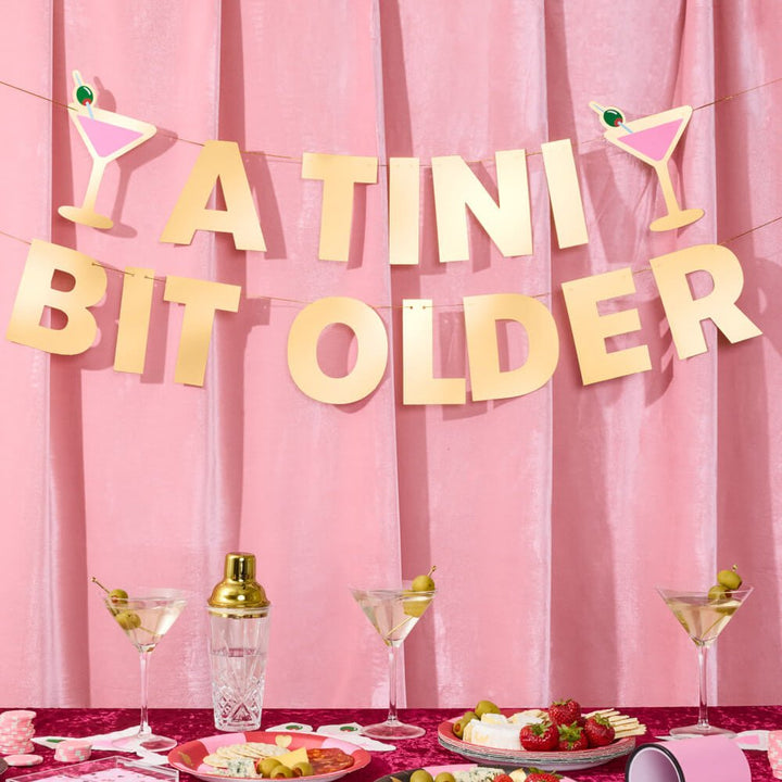 A Tini Bit Older banner hung on pink velvet drapes above a martini bar with olive cocktails and a gold shaker
