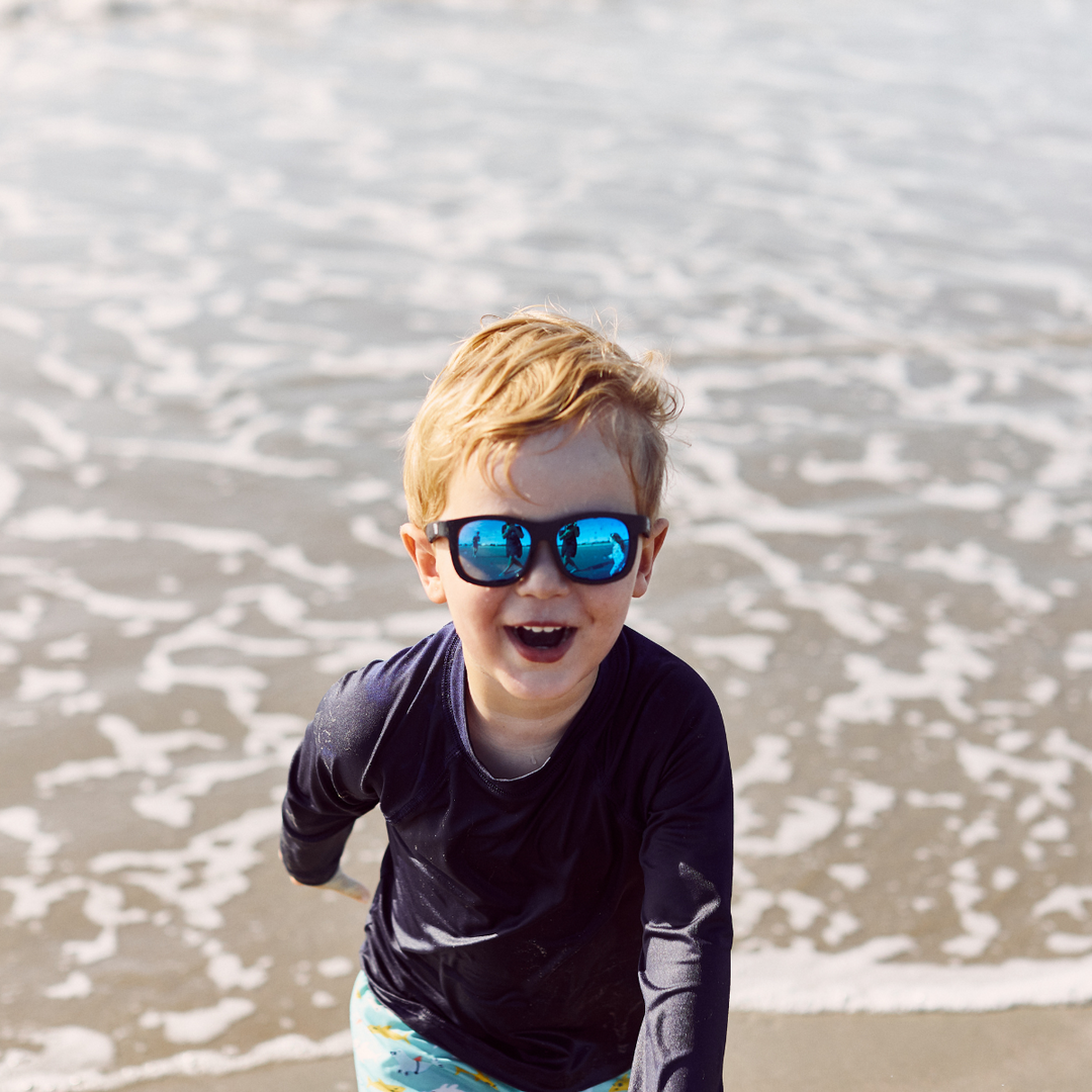 A blond boy at the beach wearing black aviator sunglasses with blue mirrored lenses, waves behind him.