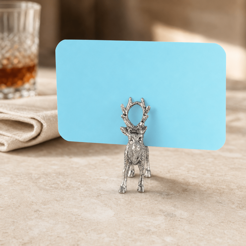 Pewter stag place card holder with a blue card on a stone table beside a glass of whiskey