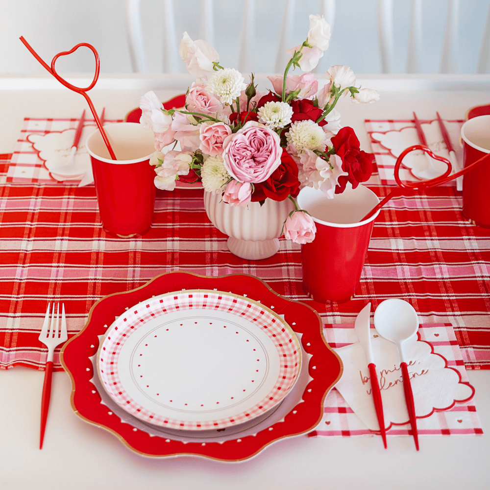 Valentine's table with red heart straws in red cups, florals, and scalloped gingham plates