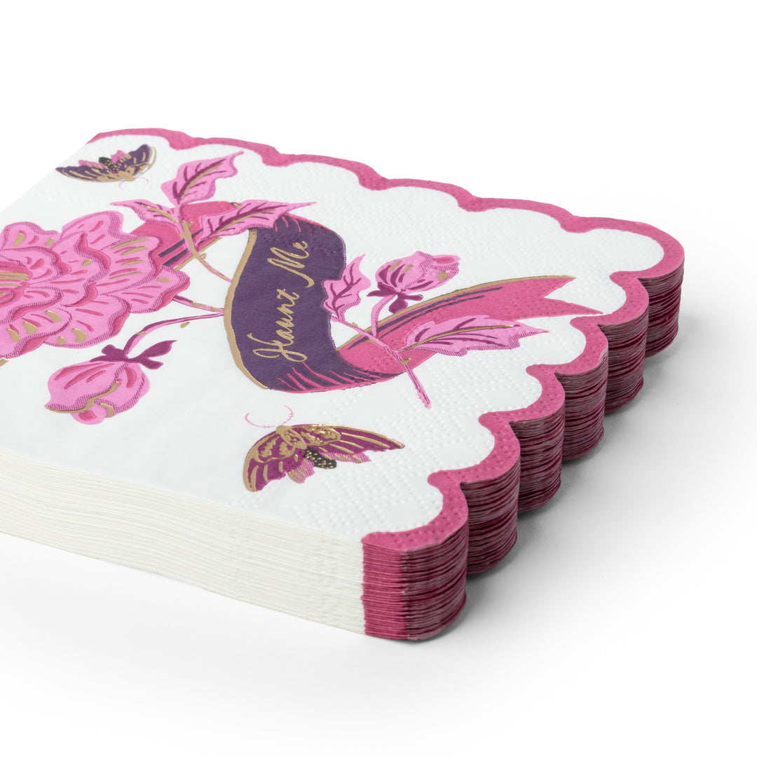 A stack of white paper napkins with scalloped edges, decorated with pink floral designs, butterflies, and a purple banner reading 'Haunt Me'.