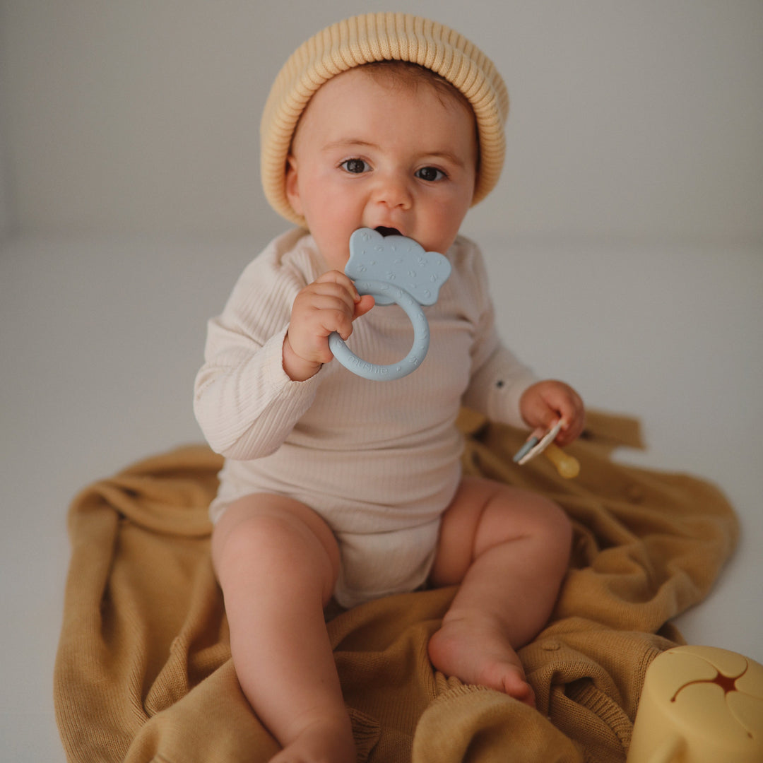 Baby in a knit hat mouthing a blue Mushie animal ring teether on a mustard blanket