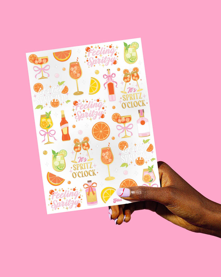 Hand holding a full sheet of spritz cocktail temporary tattoos in gold foil, pink, and orange