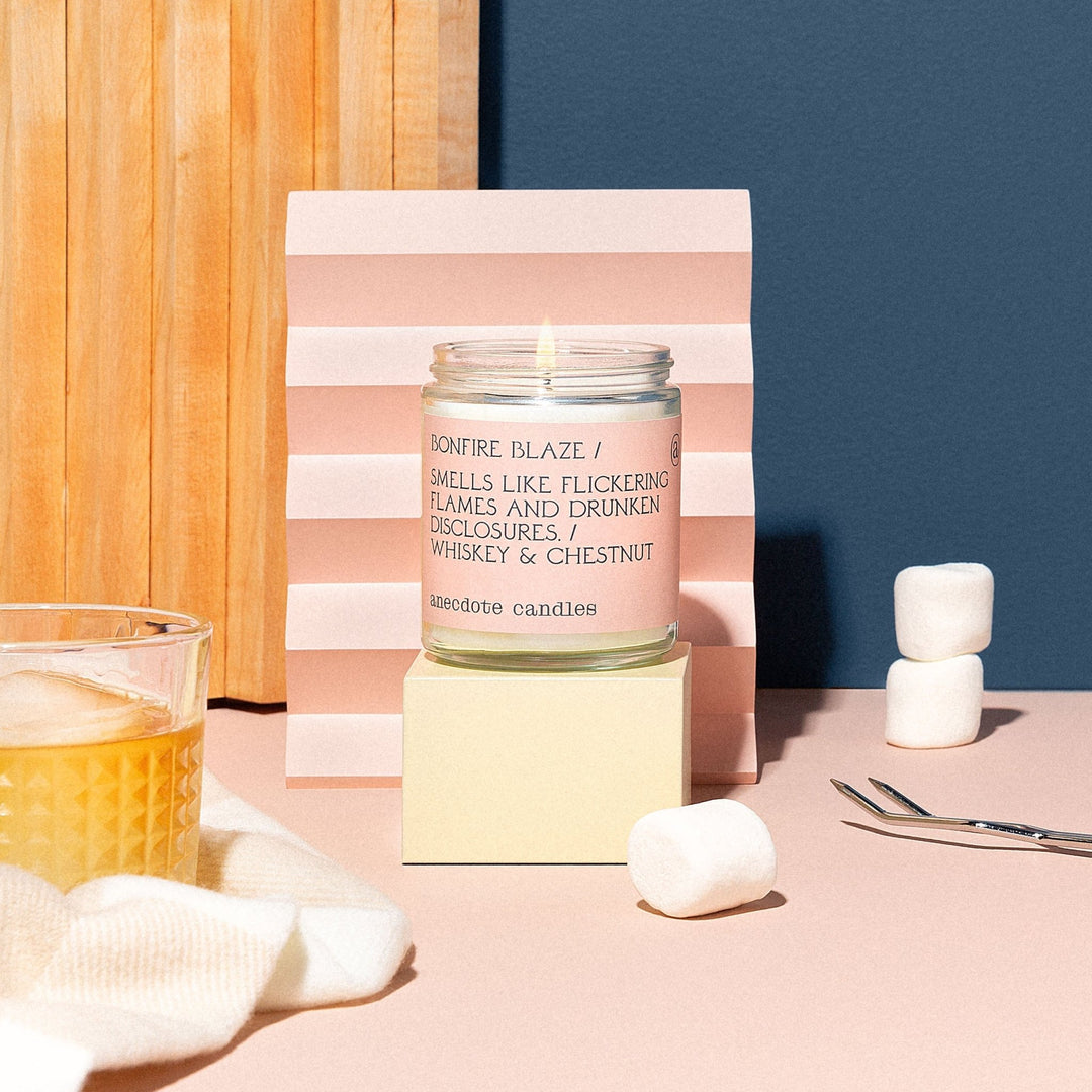 Lit Bonfire Blaze candle on a cream block with marshmallows, a whiskey glass, and a pink folded backdrop
