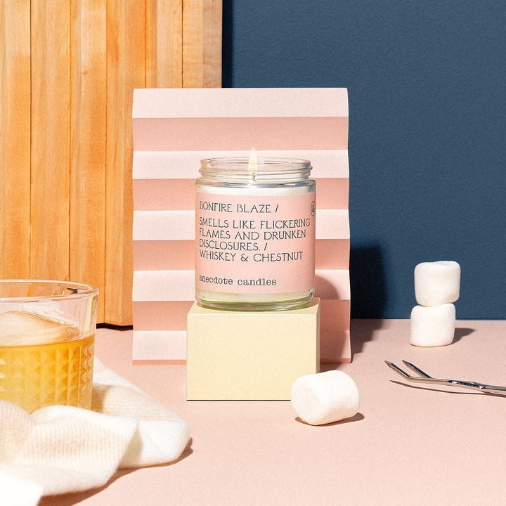 Lit Bonfire Blaze candle on a cream block with marshmallows, a whiskey glass, and a pink folded backdrop
