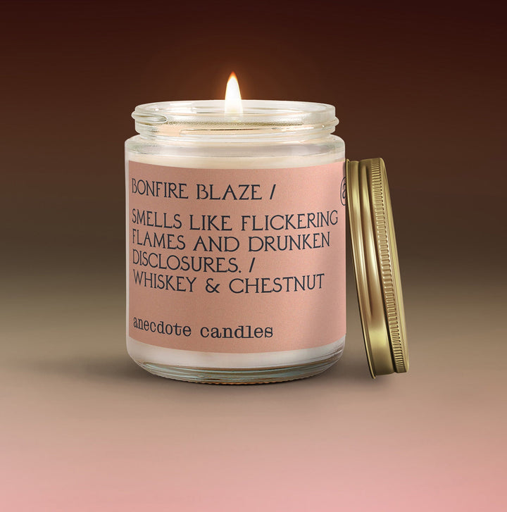 Lit Bonfire Blaze whiskey and chestnut candle with a blush pink label and gold lid leaning beside the glass jar