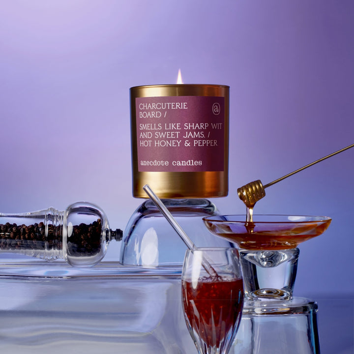 Lit Charcuterie Board candle elevated on glassware beside a honey dipper, honey, and a pepper grinder against purple