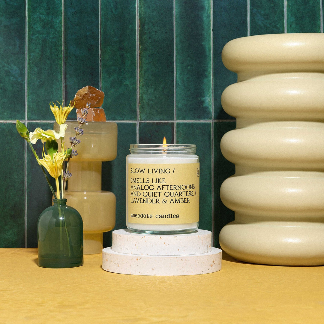 Lit Slow Living candle on stacked round plinths beside flowers and yellow ceramics against green wall tiles