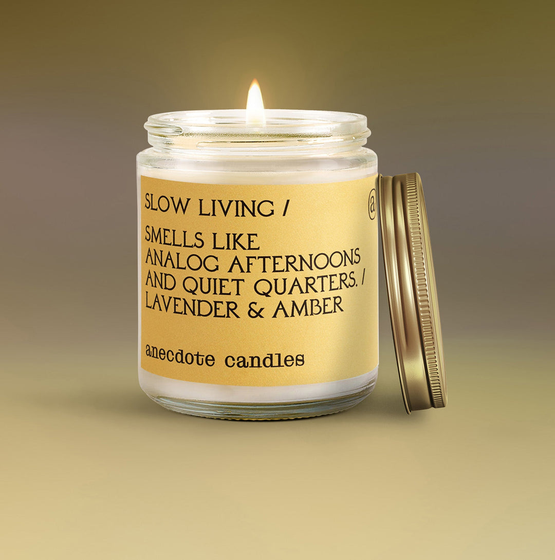 Lit Slow Living lavender and amber candle with a yellow label and gold lid leaning beside the glass jar