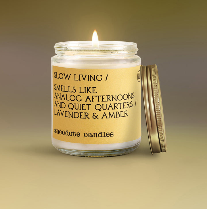 Lit Slow Living lavender and amber candle with a yellow label and gold lid leaning beside the glass jar