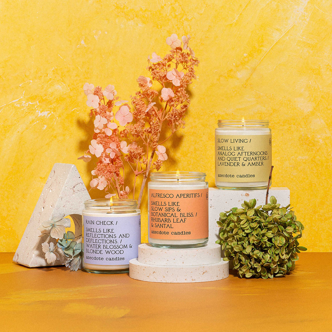 Slow Living, Alfresco Aperitifs, and Rain Check jar candles on white display stands with dried flowers against yellow