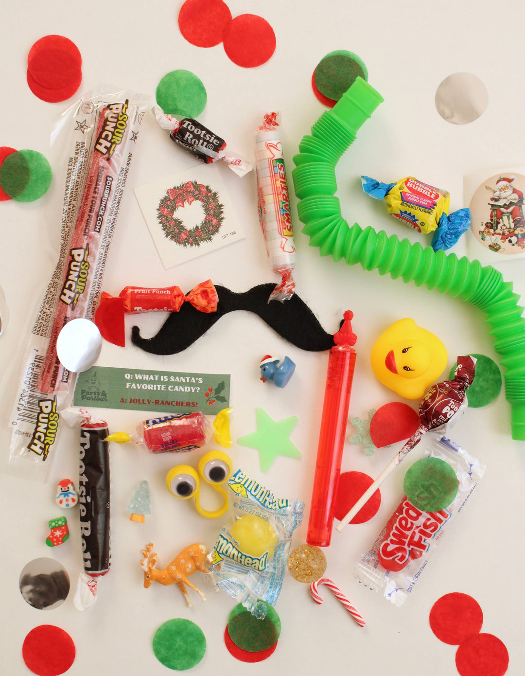 Flat lay of jumbo bon bon fillers: candies, a pop tube, a mustache, a yellow toy, and red and green confetti for Christmas
