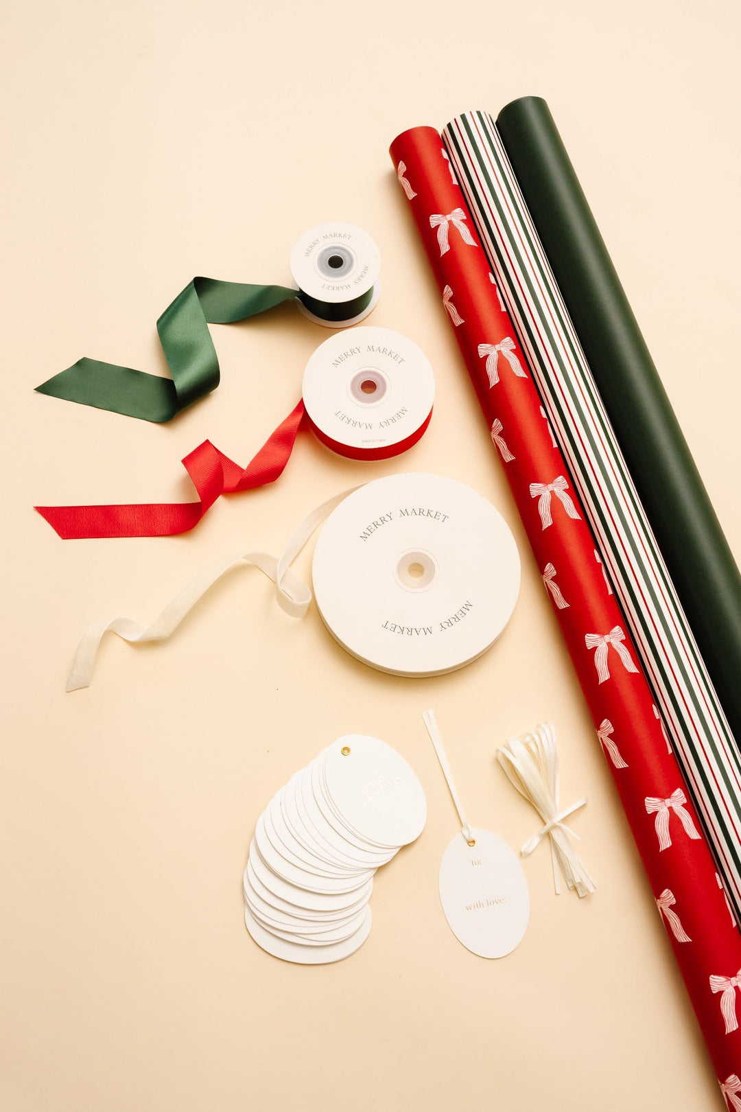 Bows Christmas wrapping paper kit flat lay: three rolls, green, red, and cream ribbons, and oval gold foil tags on cream