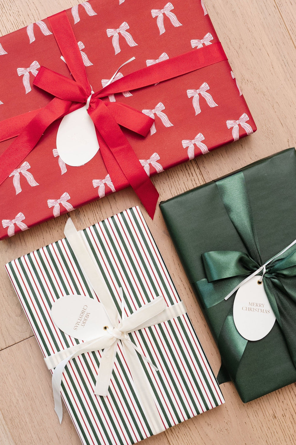Gifts wrapped in red bow print, red and green stripe, and solid green paper with satin bows on a wood floor