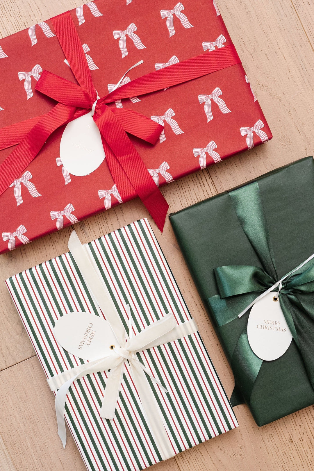 Gifts wrapped in red bow print, red and green stripe, and solid green paper with satin bows on a wood floor