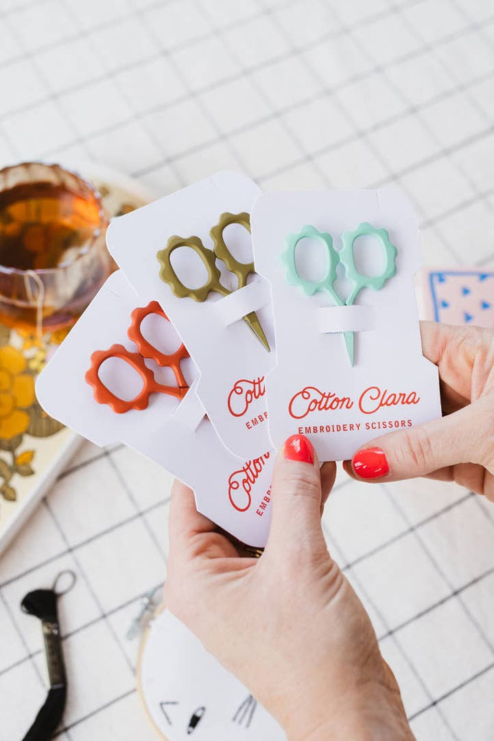 Hand holding rust, olive, and mint flower embroidery scissors cards