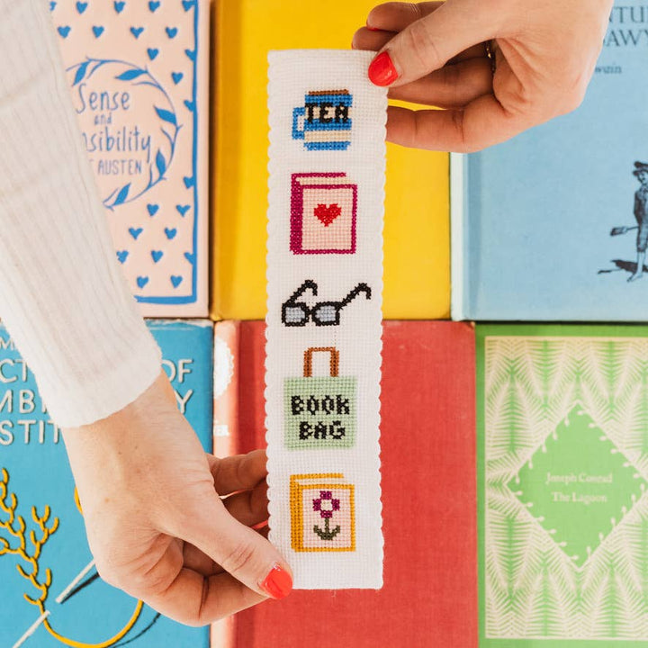 Holding the bookish cross stitch bookmark above a stack of colorful vintage books