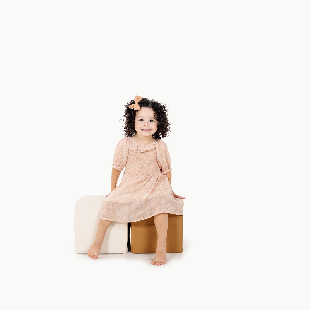 Toddler in a blush dress sitting on camel and ivory foam play cubes for active play.